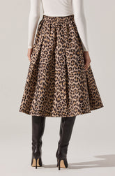A woman is wearing a high-waisted, full-length skirt with a leopard print pattern in shades of brown and black. The skirt has a slight sheen and a voluminous, A-line silhouette. She pairs it with a long-sleeved, fitted white top and black knee-high boots with high stiletto heels.